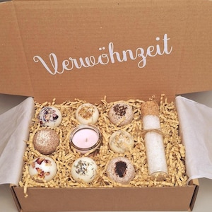 Könnte beinhalten: Ein brauner Pappkarton gefüllt mit Badebomben, einer kleinen Kerze und einem Glasröhrchen mit Badesalz. Die Badebomben sind rund und mit getrockneten Blumen und Kräutern verziert. Auf dem Karton steht das Wort "Verwöhnzeit".