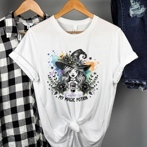 May include: White t-shirt featuring a witch with a black hat, holding a coffee cup, surrounded by floral and watercolor elements. The text "Coffee is my magic potion" is printed below the image.