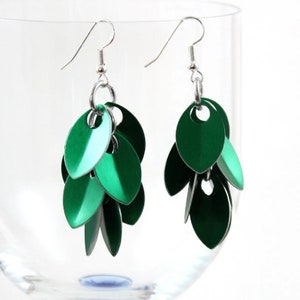 May include: A pair of dangle earrings featuring a cluster of green metal leaves. The earrings are made from silver metal and have a hook fastening.