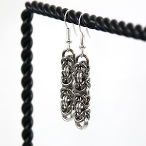 May include: A pair of silver chain earrings with a dangling design. The earrings are hanging from a black stand.