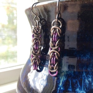 May include: A pair of silver earrings with purple accents. The earrings are made of chainmail and hang from a silver hook.