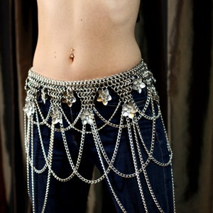 May include: A silver chainmail belt with flower-shaped charms hangs from the waist and extends down the legs. The belt is worn over blue jeans.