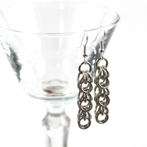 May include: Silver chain earrings with a delicate, linked design. The earrings are hanging from a glass goblet.