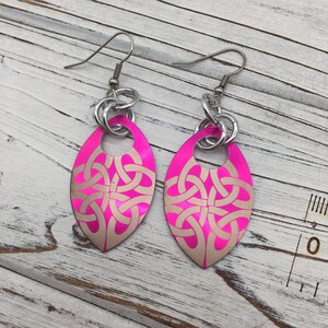 Peut inclure: Une paire de boucles d'oreilles ovales de couleur rose et argent avec un motif de nœud celtique. Les boucles d'oreilles ont une boucle argentée en haut.