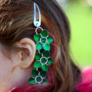 May include: A silver hair clip with a chain of green metal flowers. The flowers are made of small metal pieces that are linked together.
