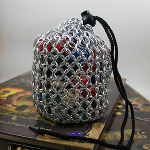 Peut inclure: Un sac à dés en maille de chaîne argentée avec un cordon de serrage noir. Le sac est rempli de dés colorés.