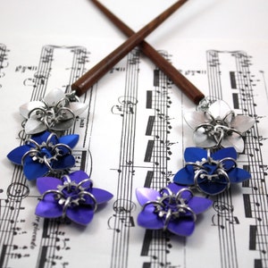 May include: A pair of chopsticks with silver and blue flower-shaped hair clips. The chopsticks are made of wood and have a dark brown finish. The hair clips are made of metal and have a shiny finish. The hair clips are arranged in a row on a sheet of music.