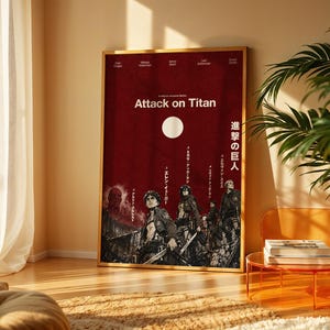 May include: A rectangular poster in a gold frame featuring a deep red background with the text Attack on Titan. The artwork displays several characters in dark attire, a large white circle, and Japanese text. The poster is set against a wall.