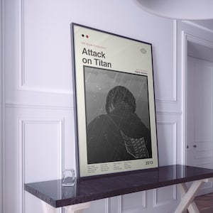 Op de afbeelding: Een ingelijste poster met de titel "Attack on Titan". De poster heeft een minimalistisch ontwerp met een grijswaarden illustratie van een persoon in een jas. De poster wordt tentoongesteld op een donkere tafel tegen een witte muur.