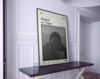 May include: A framed poster featuring the text "Attack on Titan" in a minimalist design. The poster's central image is a grayscale illustration of a person in a jacket. The poster is displayed on a dark table against a white wall.