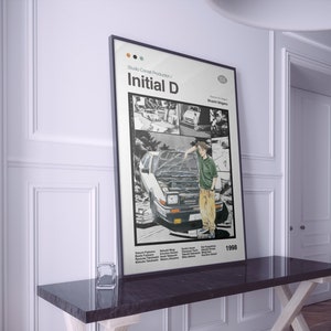 Puede incluir: Un póster en blanco y negro con fondo blanco que presenta una ilustración manga de un coche y un joven. El póster tiene el texto "Studio Comet Production / Initial D" y "Shuuichi Shigeno" en texto negro. El póster también tiene el año "1998" en texto negro.