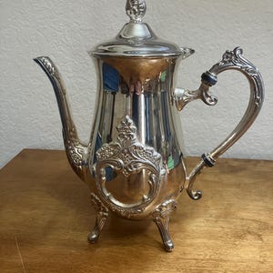 May include: A silver-colored tea or coffee pot with ornate detailing. The pot features a curved spout, a decorative handle, and a lid with a finial. The body of the pot has an embossed design and rests on three small feet. The pot is likely made of metal.