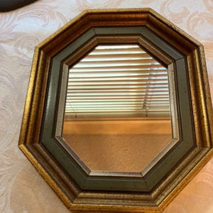 May include: An octagonal wall mirror featuring a multi-layered frame with an outer gold-toned border and an inner dark green border with subtle speckling. The reflective glass surface shows a reflection of horizontal window blinds.