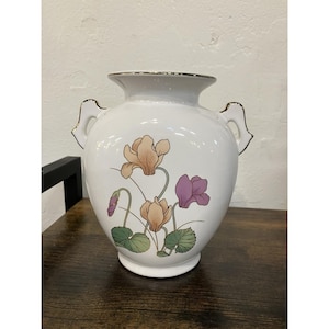 May include: White ceramic vase with two handles and a gold rim. The vase features a floral design with peach and purple flowers, green leaves, and a small purple bud. The vase is a decorative home decor item.