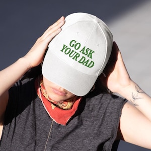 May include: A white baseball cap with the text "GO ASK YOUR DAD" in green. The cap is worn by a person, and the background is a neutral gray.