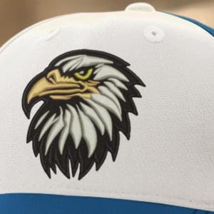 May include: White and blue trucker hat featuring an embroidered eagle head with a yellow beak and green eyes. The eagle design is detailed with black and white feathers. The hat has a white front panel and a blue mesh back.