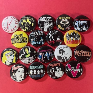 May include: A collection of round, black, white, and yellow punk rock band buttons. The buttons feature band names like "Blondie", "Sex Pistols", "The Clash", and "Ramones", along with various graphics and text. The buttons are arranged on a red surface.