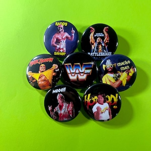 May include: A collection of seven circular pin-back badges featuring professional wrestlers. The badges display images of Randy Savage, Stone Cold Steve Austin, Hulk Hogan, the WWF logo, Macho Man, Ric Flair, and Roddy Piper with their names.