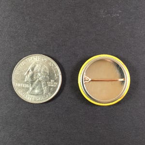 Puede incluir: Una moneda de veinticinco centavos estadounidense junto a un bot&oacute;n amarillo con un alfiler de seguridad. El bot&oacute;n tiene un borde amarillo y un interior plateado. La moneda muestra el perfil de George Washington.
