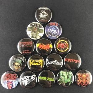 May include: A collection of black metal band button pins. The pins feature band logos and artwork, including "Onslaught," "S.O.D.," "D.R.I.," "Slayer," and "Exciter." The pins are round and have a glossy finish.