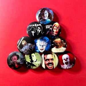 Puede incluir: Una colección de diez chapas redondas dispuestas en forma de pirámide. Cada chapa presenta una colorida ilustración gráfica de un famoso personaje de película de terror, incluyendo figuras de Scream, Child's Play y otras películas clásicas.