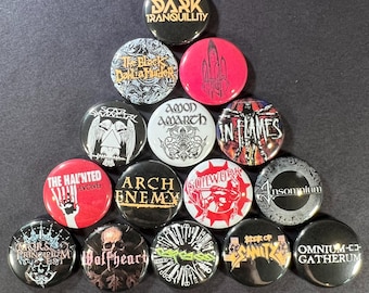 May include: A collection of round metal pinback buttons featuring various band logos and names. The buttons are in a variety of colors, including black, red, white, and gold. The text on the buttons includes band names like "Dark Tranquillity" and "Arch Enemy".