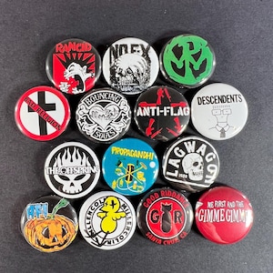 May include: A collection of round pinback buttons featuring various band logos and graphics. Designs include skulls, text, and symbols in red, black, white, and green. The buttons are arranged on a dark surface.