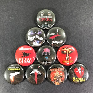 Puede incluir: Un conjunto de botones redondos con títulos de películas e imágenes. Los botones son rojos, negros y blancos, con textos como "Opera", "Phenomena" y "Suspiria". Los botones están dispuestos sobre una superficie oscura.