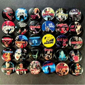 Puede incluir: Una colección de 30 botones redondos y negros con títulos e imágenes de películas. Los títulos incluyen "The Thing", "Child's Play", "Evil Dead" y "Halloween II". Los botones muestran varios temas y diseños de películas de terror.