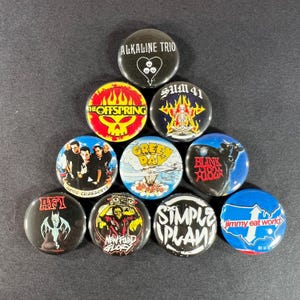 May include: A collection of ten round pinback buttons featuring band logos and names. The buttons display various colors and designs, including text and graphics from bands like Alkaline Trio, Sum 41, and Blink 182.