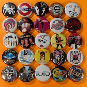 May include: A collection of 25 round pinback buttons featuring various band logos and portraits. Bands include Genesis, The Police, Billy Joel, Elvis Costello, and more. The buttons are arranged on an orange surface.