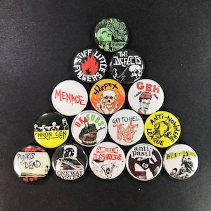 May include: A collection of punk rock button badges with various band names and graphics. The badges feature bold text and imagery in colors like red, black, white, and yellow. The badges are arranged on a dark surface.