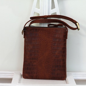 Könnte beinhalten: Eine braune Leder-Umhängetasche mit einer Kroko-Prägung. Die Tasche hat einen Klappverschluss und einen langen, verstellbaren Riemen.