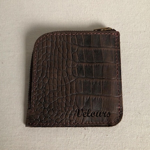 Peut inclure: Un portefeuille marron texturé avec une fermeture éclair. Le portefeuille présente un motif peau de crocodile et le mot "Velours" est imprimé en bas à droite. La fermeture éclair est dorée et les coutures sont rouges.