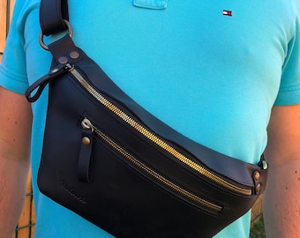 May include: Black leather crossbody bag with gold-colored zippers and hardware. The bag has a long adjustable strap and a curved design. The bag is worn across the body. The bag has the word "Delores" embossed on the front.