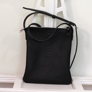 May include: Black crossbody bag with a snakeskin texture. The bag has a long, adjustable strap and a shorter handle. The zipper closure is silver. The bag is sitting on a white surface.