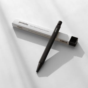 Può includere: Una penna nera con la scritta "prototo" stampata sul lato, appoggiata su una scatola bianca. La scatola presenta anche il testo "Zero Waste Kugelschreiber". La penna ha un design geometrico.