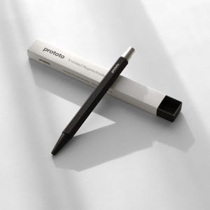 Peut inclure: Un stylo noir au design géométrique et à la pointe argentée, à côté de sa boîte blanche. Le mot "prototo" est imprimé sur le stylo et la boîte. La boîte porte également l'inscription "Kreislauf Kugelschreiber".