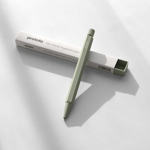 Op de afbeelding: Een lichtgroene pen met een zeshoekig ontwerp, naast de witte doos. De doos en pen zijn voorzien van de tekst "prototo" en "Zero Waste Kugelschreiber". De pen staat schuin en werpt een schaduw op het witte oppervlak.
