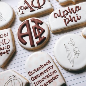 May include: Assortment of decorated sugar cookies with white icing. Some cookies feature sorority letters and phrases like "Union Hand in Hand" and "Sisterhood Generosity Innovation Character" in brown icing. Other cookies have floral designs.