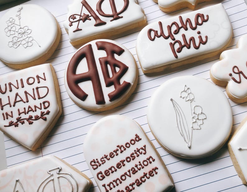 May include: Assortment of decorated sugar cookies with white icing. Some cookies feature sorority letters and phrases like "Union Hand in Hand" and "Sisterhood Generosity Innovation Character" in brown icing. Other cookies have floral designs.