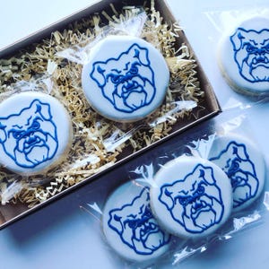 May include: Round sugar cookies with white icing and a blue bulldog design. Some cookies are packaged in clear plastic, while others are in a brown box with shredded paper. The cookies are a light beige color.