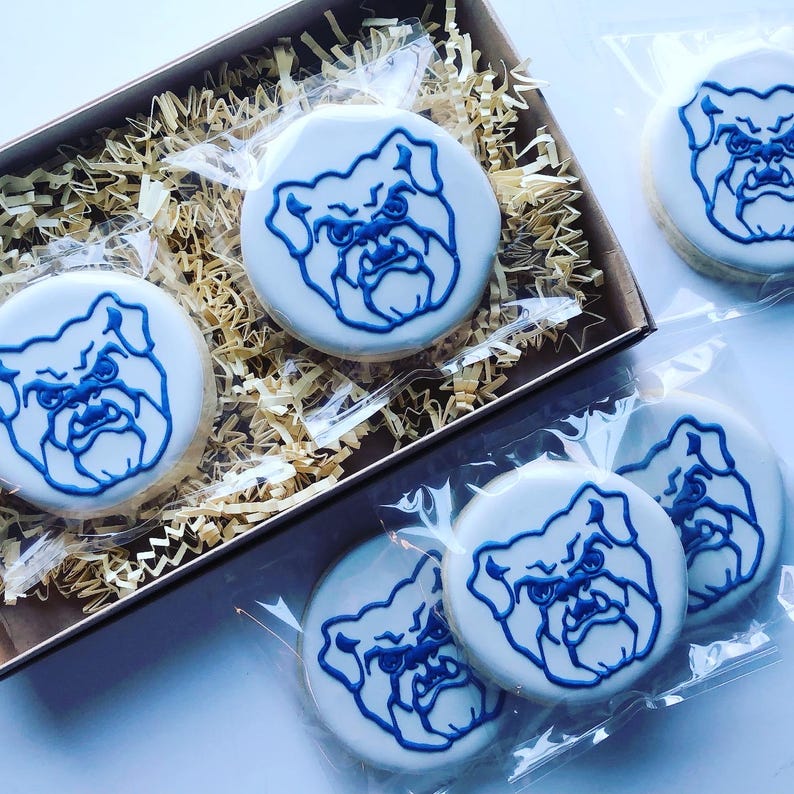 May include: Round sugar cookies with white icing and a blue bulldog design. Some cookies are packaged in clear plastic, while others are in a brown box with shredded paper. The cookies are a light beige color.