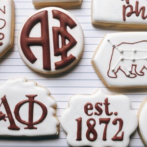 May include: Assortment of decorated sugar cookies with white icing and brown designs. Some cookies feature Greek letters, a bear constellation, and the text "UNION HAND IN HAND" and "est. 1872".