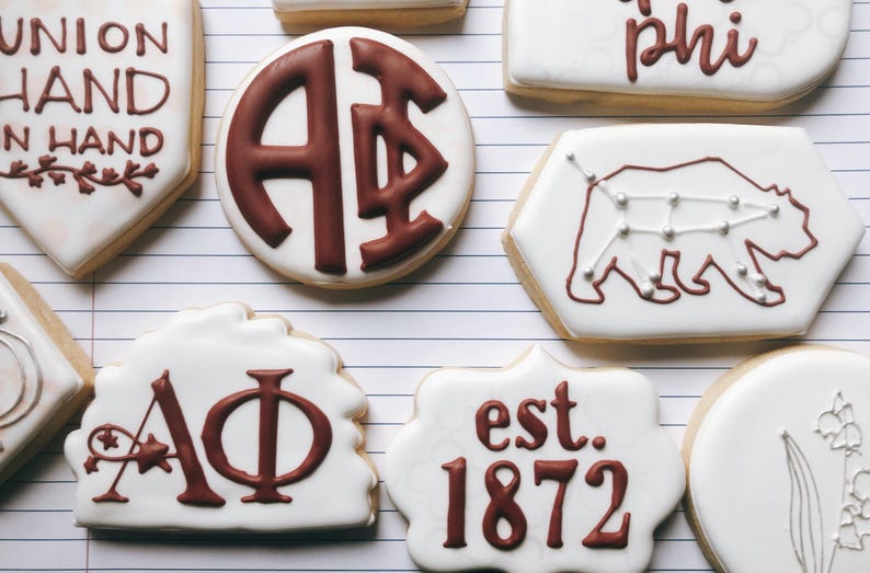 May include: Assortment of decorated sugar cookies with white icing and brown designs. Some cookies feature Greek letters, a bear constellation, and the text "UNION HAND IN HAND" and "est. 1872".