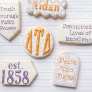 May include: Assortment of decorated cookies with various designs and text. One cookie reads "Truth, Courage, Faith, Power." Another says "Aidan." Other cookies feature "Delta Tau Delta" and "Committed to Lives of Excellence."