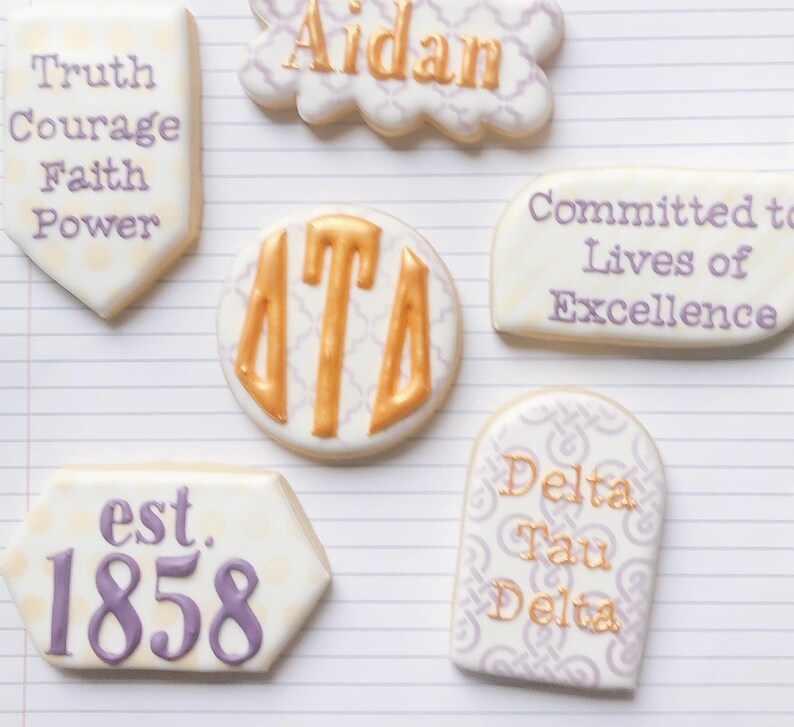 May include: Assortment of decorated cookies with various designs and text. One cookie reads "Truth, Courage, Faith, Power." Another says "Aidan." Other cookies feature "Delta Tau Delta" and "Committed to Lives of Excellence."