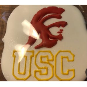 May include: A white frosted cookie with a red and yellow design. The red design is a stylized symbol, and the yellow letters "USC" are below it. The cookie has a glittery finish.