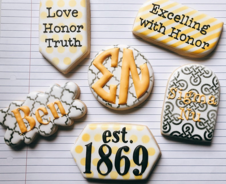 May include: Assortment of decorated cookies with various designs and text. One cookie reads "Love Honor Truth", another "Excelling with Honor". Other cookies feature "Ben", "Sigma Nu", and "est. 1869". The cookies have white, yellow, and black icing.
