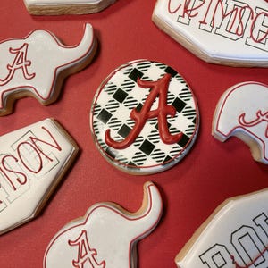 May include: Assortment of decorated sugar cookies on a red surface. The cookies feature white icing with red accents, including the letter "A" and team names. Some cookies are elephant-shaped, while others are rectangular or round.